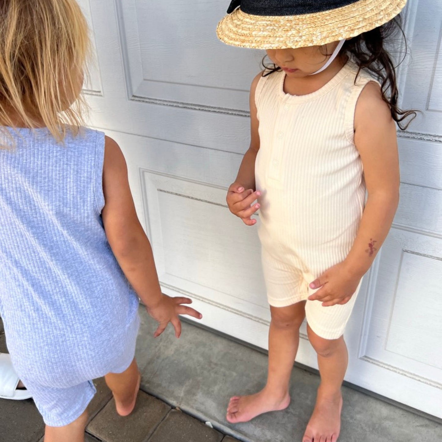 Two children standing on a step, one in a gray jumpsuit and the other in a vanilla sleeveless outfit with a straw hat.